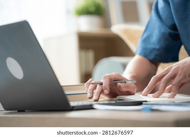 Close up of a man hand using a laptop trackpad and holding a pen over a notebook on the table, representing remote working, online learning, and office business