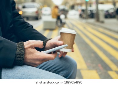 Close up of man hands holding mobile phone, using internet while sitting on the bench and drinking take away cup of coffee. Chatting with friends or family, reading latest news and updates in the web. - Shutterstock ID 1897054990
