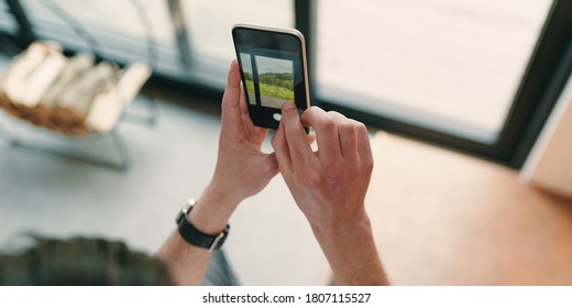 Close Up Of Man Hands Taking Picture Of The Beautiful View Outside The Window With Mobile Phone. Businessman Using Smartphone In His Modern House To Photograph The Mountains Through The Window