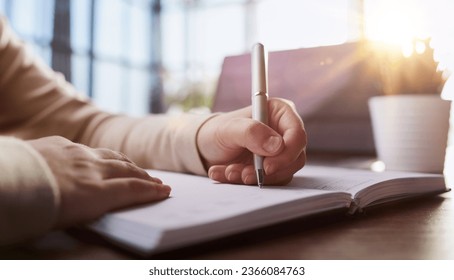 Close up of man hands writing in spiral notepad placed on wooden desktop with various items