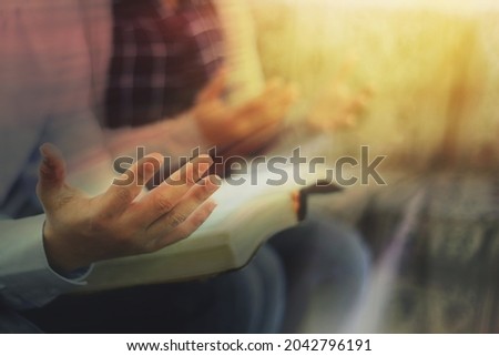 Close up of a man holding a bible and raise hand up praying to God with his friends while sitting on the sofa at home with window light rays, Christian family worship concept, copy space 