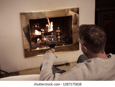 Close Up Of Man Holding And Drinking Red Wine Glass By Open Wood Fire Place Relaxing And Enjoying Quiet Moment At Home In Winter Holiday Vacation, Christmas Or Leisure Concept.