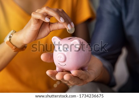 Close up of man holding pink piggybank while woman putting coin in it. Indian young couple saving money for their wedding. Close up of woman hand putting euro money in piggy bank to save.