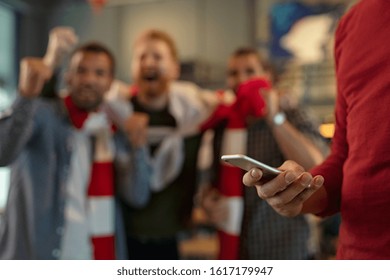 Close up of man holding smartphone and betting on the game while supporters celebrating the victory in background. Soccer fans cheering for league in pub, man using smart phone to gamble, copy space. - Powered by Shutterstock