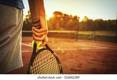 Close up of man holding tennis racket on clay court. In his hand is tennis ball. On court is sunset./Man holding tennis racket