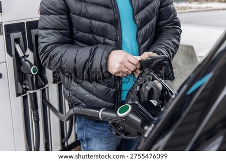 Close up of man holding wallet while refueling car at gas station. Concept of paying for fuel, gasoline prices, transportation cost and everyday expenses.