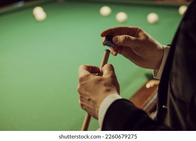 Close up of man in a jacket rubs the end of a cue with chalk before the game. Background of green pool table with billiard balls