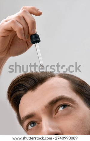 Close up of man looking up while applying liquid serum with dropper to hair on light gray background in minimal studio setting.