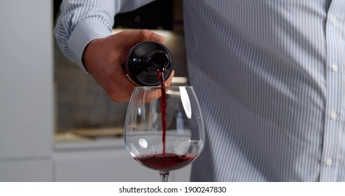 Close Up Of Man Pouring Glass Of Red Wine In Kitchen At Home. Guy In Shirt Filling Glass With Wine Standing Alone In Modern Kitchen And Having Dinner