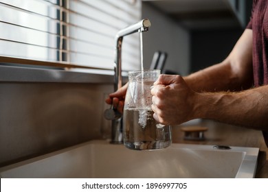 Close up of a man pouring water in the jug from a tap while standing at the kitchen sink