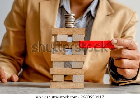 Close Up Man Pulls Red Block From Wooden Tower Stacked With Coins, Symbolizing Financial Risk