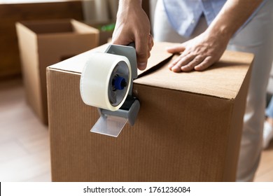 Close up man sealing cardboard box with adhesive tape, using dispenser, moving day and relocating delivery service concept, young male preparing to relocation, packing belongings, parcel