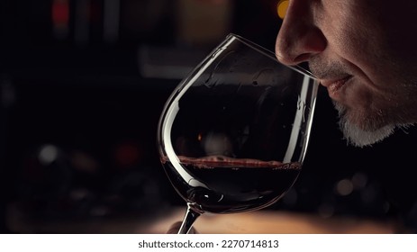 Close up man smelling red wine in wine glass. Wine expert tasting, rating and drinking wine, bottles in background.