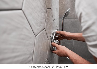 Close up of man standing by the wall with ceramic tile and installing shower faucet with metal handle in apartment. Male plumber working on bathroom renovation at home. Plumbing works concept.