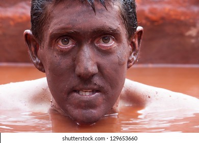 Close Up Of A Man Submerged In The Red Mud Bath With His Face Completely Covered In Mud.