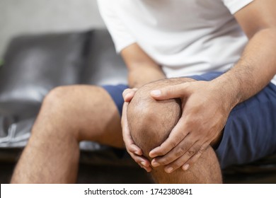 close up man suffering from feeling knee pain sitting sofa at home. hand massaging his painful knee. Health care and medical concept.
