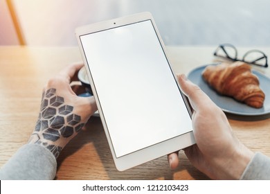 Close up of man tattooed hands holding tablet with blank screen. Man sitting behind wooden table in cafe.