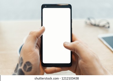 Close up of man tattooed hands holding mobile phone with blank screen. Man sitting behind wooden table in cafe.