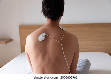 Close Up Of A Man Using A Muscle Stimulator Machine On His Back And Neck Area Using Two Electrode Pads, Sitting On The Bed In Crossed Leg Position. White And Neutral Background.