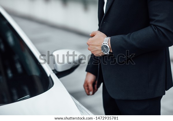 Close up of a man with a watch in his\
hand in a business suit near a premium white\
car.