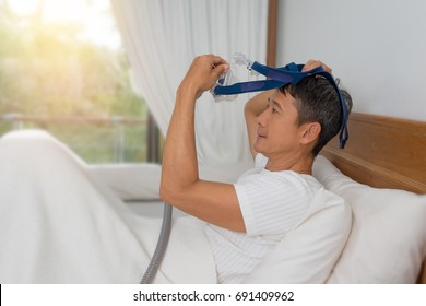 
Close Up Of Man In White Clothes Laying With Knees Up In Bed  Hands Holding And Wearing CPAP Mask . Obstructive Sleep Apnea Therapy, Side View.