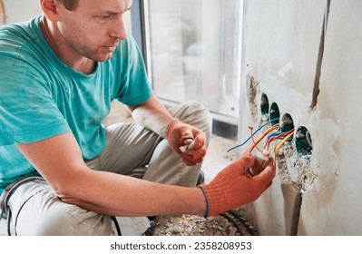 Close up of man in work gloves sitting by the wall and putting electrical wire cable in bottle with liquid. Male electrician installing electrical wiring in apartment under renovation. - Powered by Shutterstock