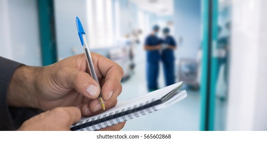 Close Up Of Man Writing In Spiral Book Against Doctor Standing On Corridor With Open Door