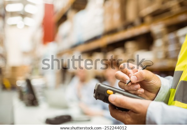 Close up of manager wearing yellow vest using\
handheld in a large\
warehouse