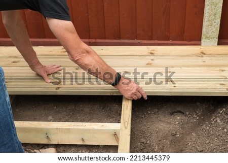 close up of  a man's arms, building a wooden decking platform in a garden against a fence.  The garden is a domestic piece of ground outside a home in autumn