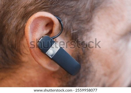A close up of a man's ear with a black Bluetooth headset attached. Texture and facial hair are detailed, with slight redness around the ear. The focus is sharp, showing the headset's sleek design.