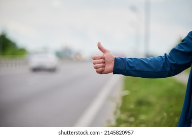 Close up of man's hand hitchhiking by roadside. Male hand showing thumbs up gesture outdoors on blurred background. Hitchhiking, hitching, auto stop concept.