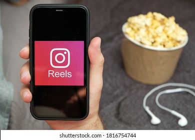 Close Up Mans Hand Holding A Mobile Phone With Instagram Reels Logo With Apple Earphones And Popcorn Box Next To Him, August 2020, San Francisco, USA