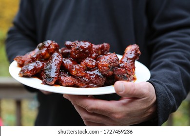 Close Up Of Man's Hand Holding A Plate Of BBQ Chicken Wings Outside.