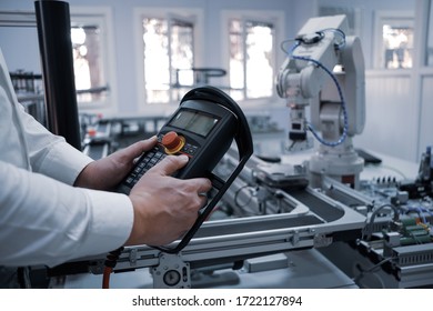 Close Up Of Man's Hand Holding Teach Pendant To Control Robot Arm Which Is Integrated On Smart Factory Production Line. Industry 4.0 Automation Line Equipped With Sensors And Robot At Background.