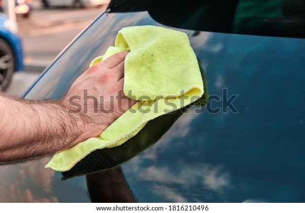 Close up mans hand polishing or cleaning a rear\
windshield of a blue automobile with a yellow microfiber rag .\
Manual car washing. 
