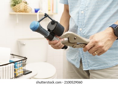 Close Up Of Man's Hands Hold Lock Jaw Pliers And Old Broken Fill Valve To Fix And Replace With Clean White Toilet And Bathroom In Background. DIY Plumbing, Home Maintenance, Handyman And House Improve