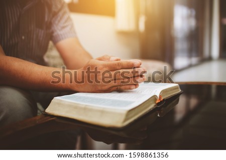 Close up of a man's hands praying on open bible while sitting on sofa at home
