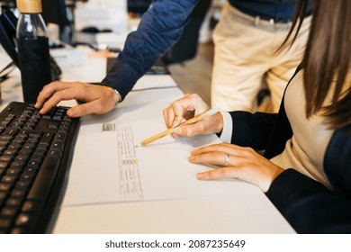 Close up of man's hands using his colleague's computer and woman's hand holding a pen, on a messy desk, in office. Working toghether