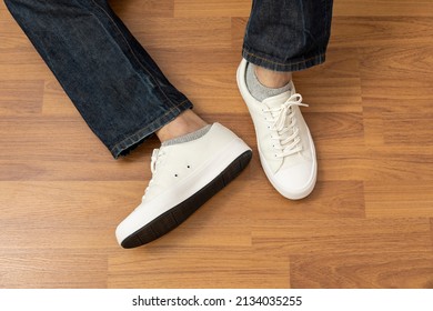 Close Up Of Man's Leg Wearing Brand New White Sneaker Shoe And Blue Jean Sitting On Wood Floor.