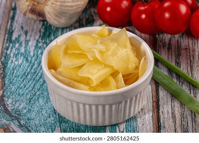 Close up of marinated ginger slices in ceramic bowl. Fresh ingredients like garlic, cherry tomatoes, and green onions surround it on rustic blue wooden table. Appetizing food still life