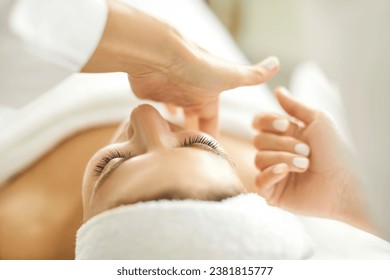 Close up masseur hands making face relaxing massage to a young girl lying with closed eyes in beauty salon or cosmetology cabinet. Professional cosmetologist making massage for woman in spa.