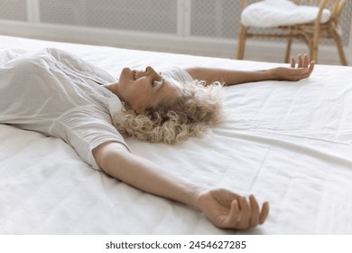 Close up mature 55s Caucasian woman falling down on white bedsheets with arms raised, take break, relaxing, enjoy fresh sheets and comfortable orthopaedic mattress for good quality sleep in bedroom - Powered by Shutterstock