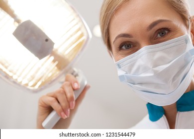 Close up of a mature female dentist smiling, wearing medical mask, adjusting dental lamp. Professional dentist preparing for teeth examination, looking to the camera. POV of patient. Medicine concept