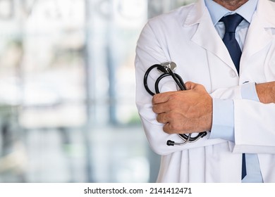 Close Up Of Mature Male Doctor With Arms Crossed Holding Stethoscope In Modern Hospital. Detail Of Mid Adult Physician Wearing Lab Coat With Folded Hands In Clinic. Proud And Satisfied Doctor.