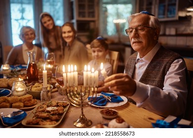 Cerca de un hombre maduro encendiendo velas en menorah mientras celebraba Hanukkah con su familia extendida a la mesa.