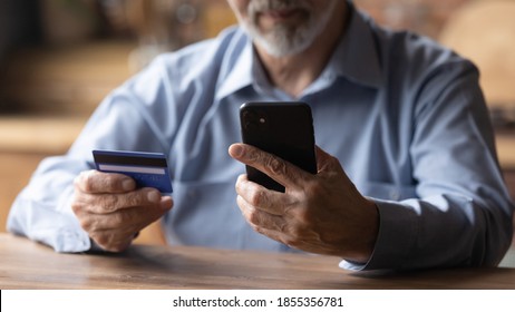 Close up mature man using phone, holding plastic credit or debit card, senior grey haired customer making secure internet payment, shopping or browsing online banking service, entering information
