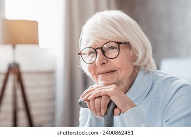 Close up of mature woman in glasses holding cane while sitting on sofa at home. Loneliness and calmness, nostalgy concept. Hospice nursing house for senior old elderly grandmother - Powered by Shutterstock