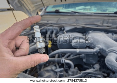 Close up of a mechanic hand holding a used spark plug near an open car engine bay during vehicle maintenance and ignition system inspection.