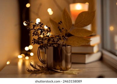 Close up of a metal copper colored cup with autumn fall leaves and dried seed branches on a wooden window sill with stacked books, lit candle and fairy lights in the blurred background - Powered by Shutterstock