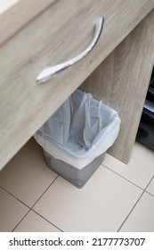 Close Up Of Metal Garbage Bin With Plastic Bag Standing Under Table. Empty Trash Can Near Workplace In Office Or Home Interior. Concept Of Maintaining Cleanliness And Waste Sorting.